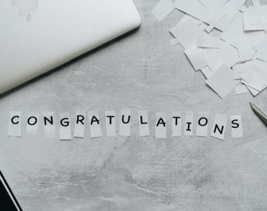 Black cut out letters on white pieces of paper laid against gray pad of paper. Photo by alleksana: https://www.pexels.com/photo/congratulations-text-in-white-pieces-of-paper-4271938/