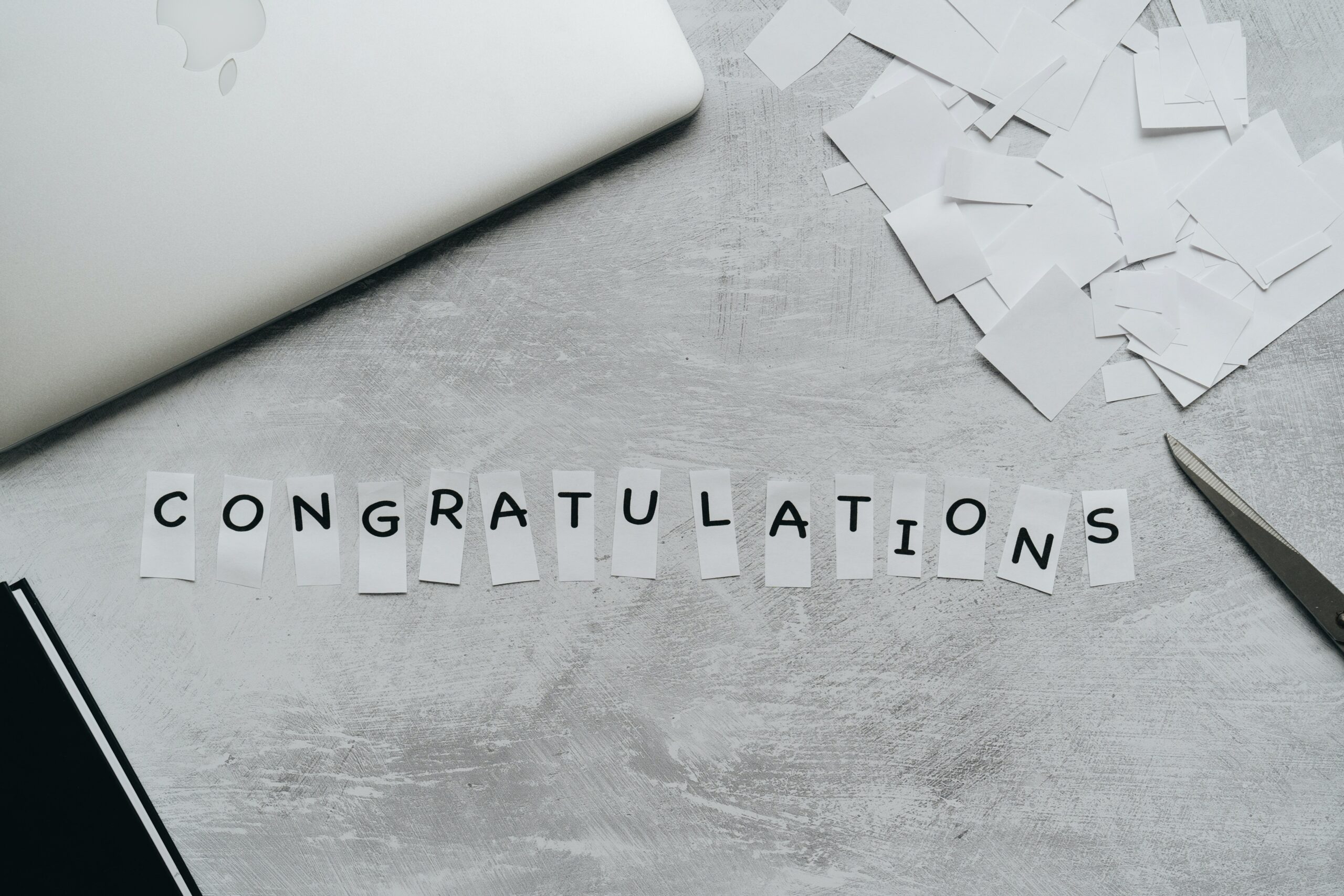 Black cut out letters on white pieces of paper laid against gray pad of paper. Photo by alleksana: https://www.pexels.com/photo/congratulations-text-in-white-pieces-of-paper-4271938/