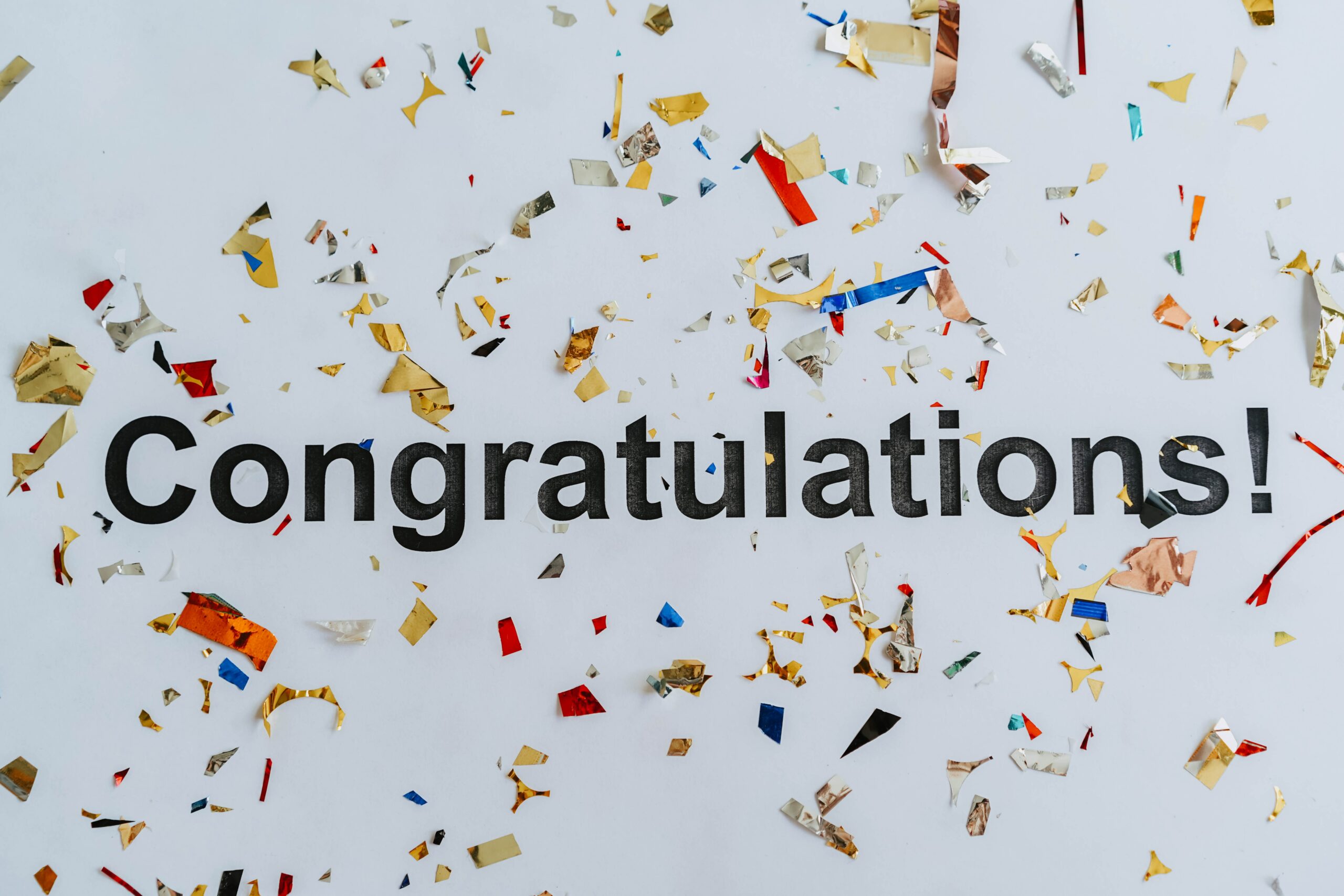 The world congratulations in bold black font in the center of a white background with multi colored confetti scattered around. Photo by olia danilevich: https://www.pexels.com/photo/congratulations-text-on-white-surface-with-confetti-8127309/