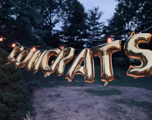 Congrats in gold lettering strung between two trees in a wooded area at dusk. Photo by Brett Garwood: https://www.pexels.com/photo/close-up-shot-of-gold-letter-balloons-6356146/