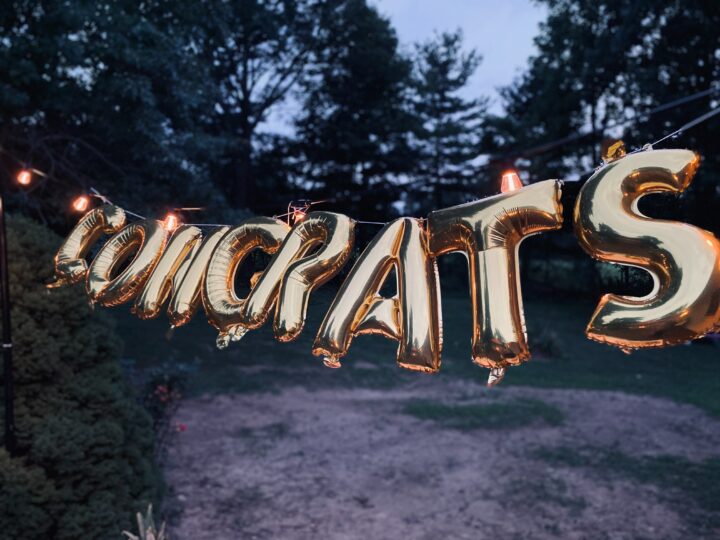 Congrats in gold lettering strung between two trees in a wooded area at dusk. Photo by Brett Garwood: https://www.pexels.com/photo/close-up-shot-of-gold-letter-balloons-6356146/